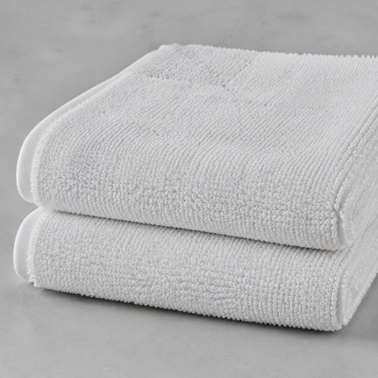 Two folded heavy white terry bath mats stacked on a pale grey surface