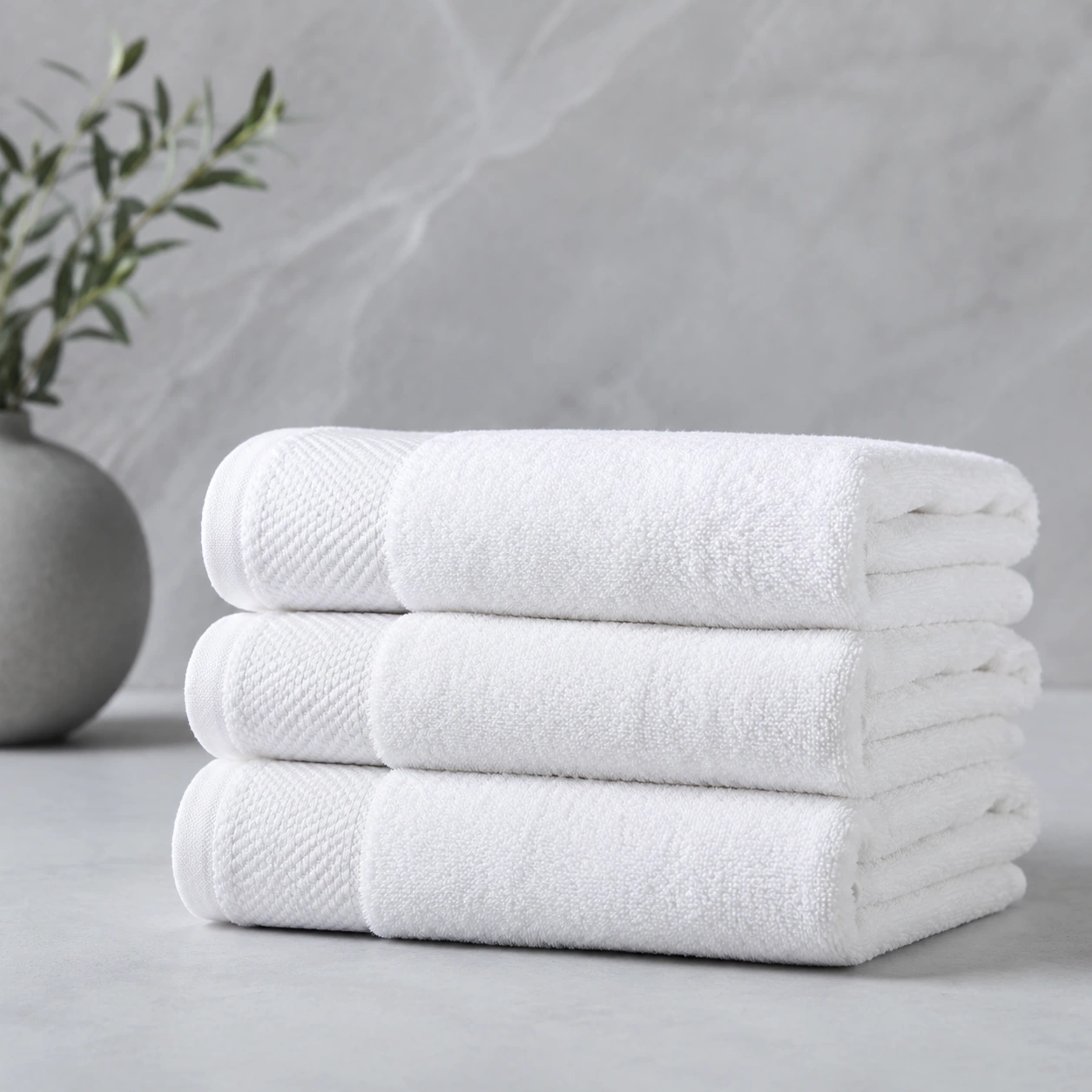 Three folded white hotel bath towels stacked on a pale grey surface