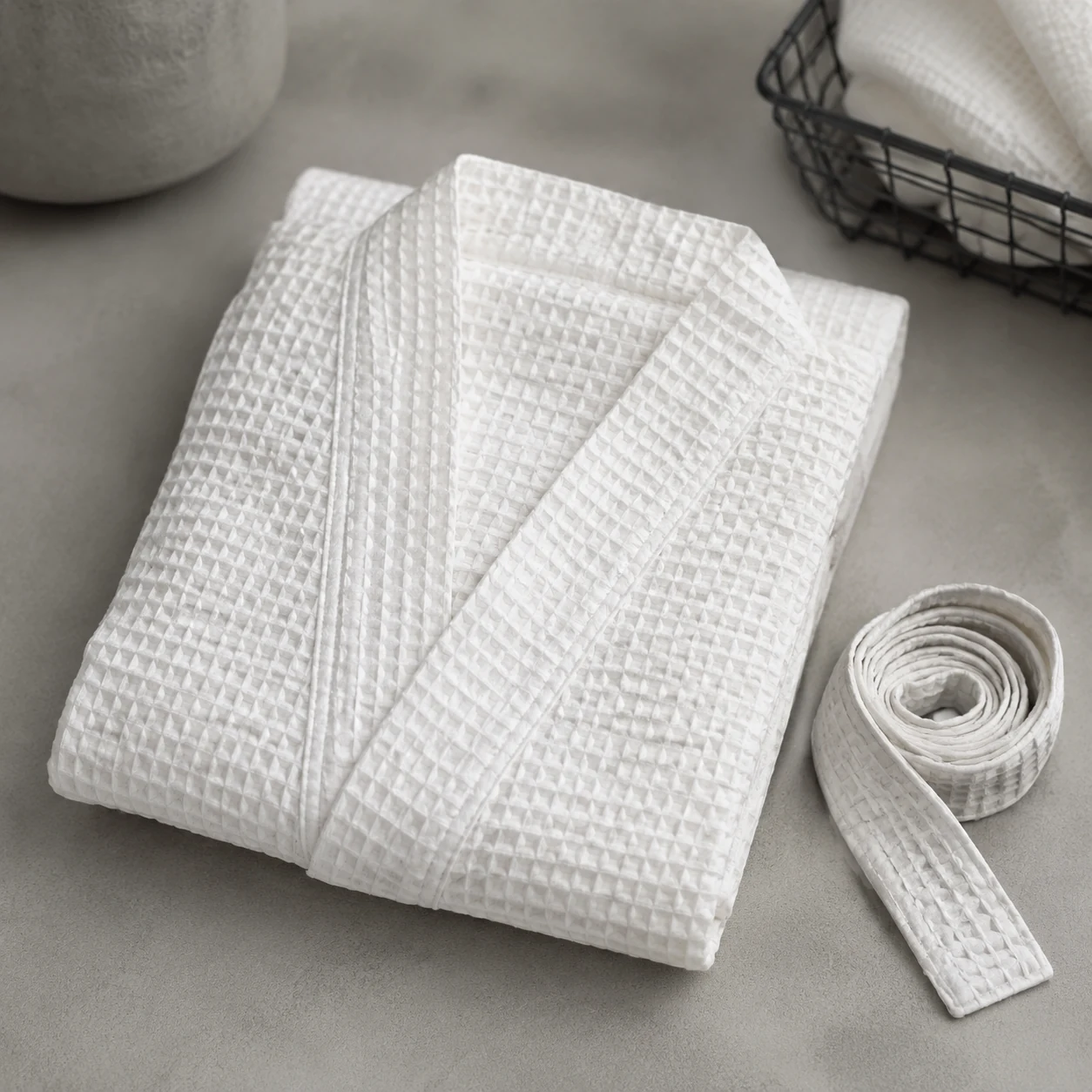 Folded white waffle weave bathrobe with a coiled belt on a pale grey surface